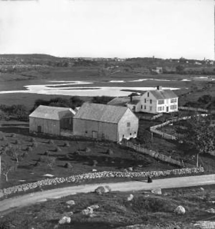 Black and white photo of a house and barns with road in foreground and pond in background.